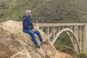 Man sitting on a rock overlooking a bridge with a mountainous landscape